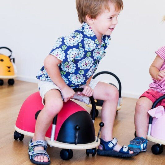 Toddler boy riding the Wheely Bug Ride On Ladybug Large ride-on toy indoors on a wooden floor