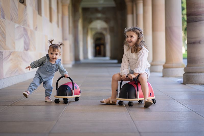 Two girls playing together with Wheely Bug Ride On Ladybug ride-on toys in an outdoor colonnade