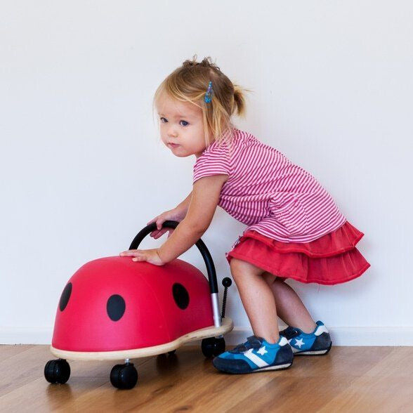 Toddler girl pushing the Wheely Bug Ride On Ladybug Large ride-on toy along a wooden floor