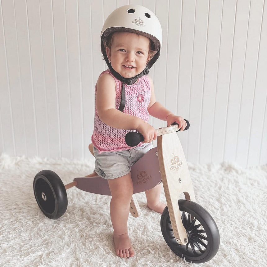 Smiling toddler in a helmet standing over the Kinderfeets Tiny Tot Plus Lavender trike