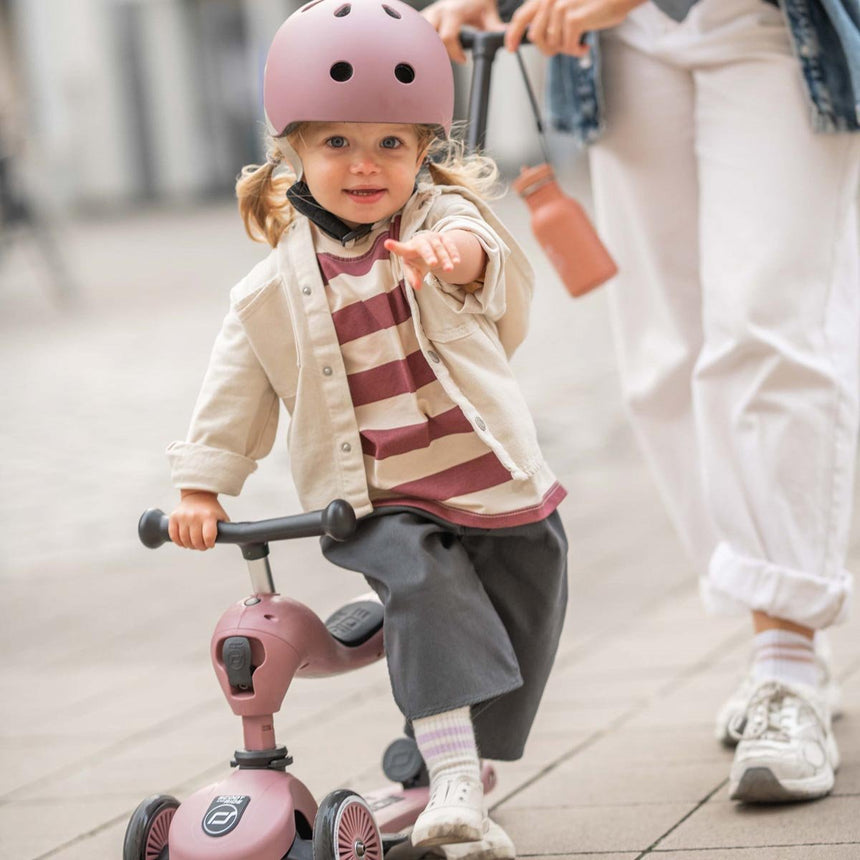Toddler wearing a Scoot & Ride Rose helmet holding a Wildberry Push & Go scooter with parent nearby