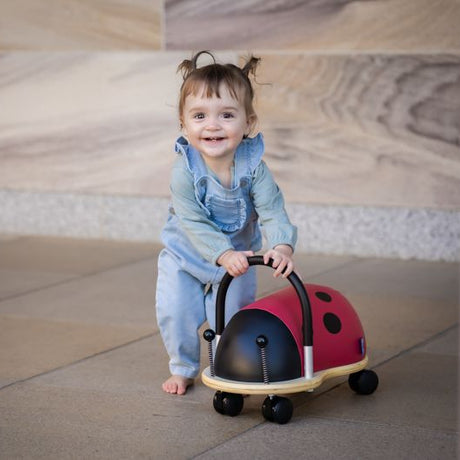 Smiling toddler girl playing with the Wheely Bug Ride On Ladybug Large ride-on toy on a tiled floor